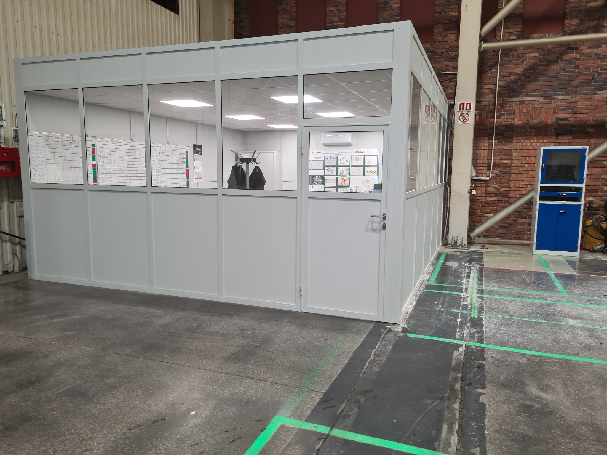 Wykończona modułowa przestrzeń biurowa w hali produkcyjnej, z widocznymi tablicami informacyjnymi i wieszakami na ubrania wewnątrz, oświetlona od góry, z zielonymi liniami wyznaczającymi strefy...