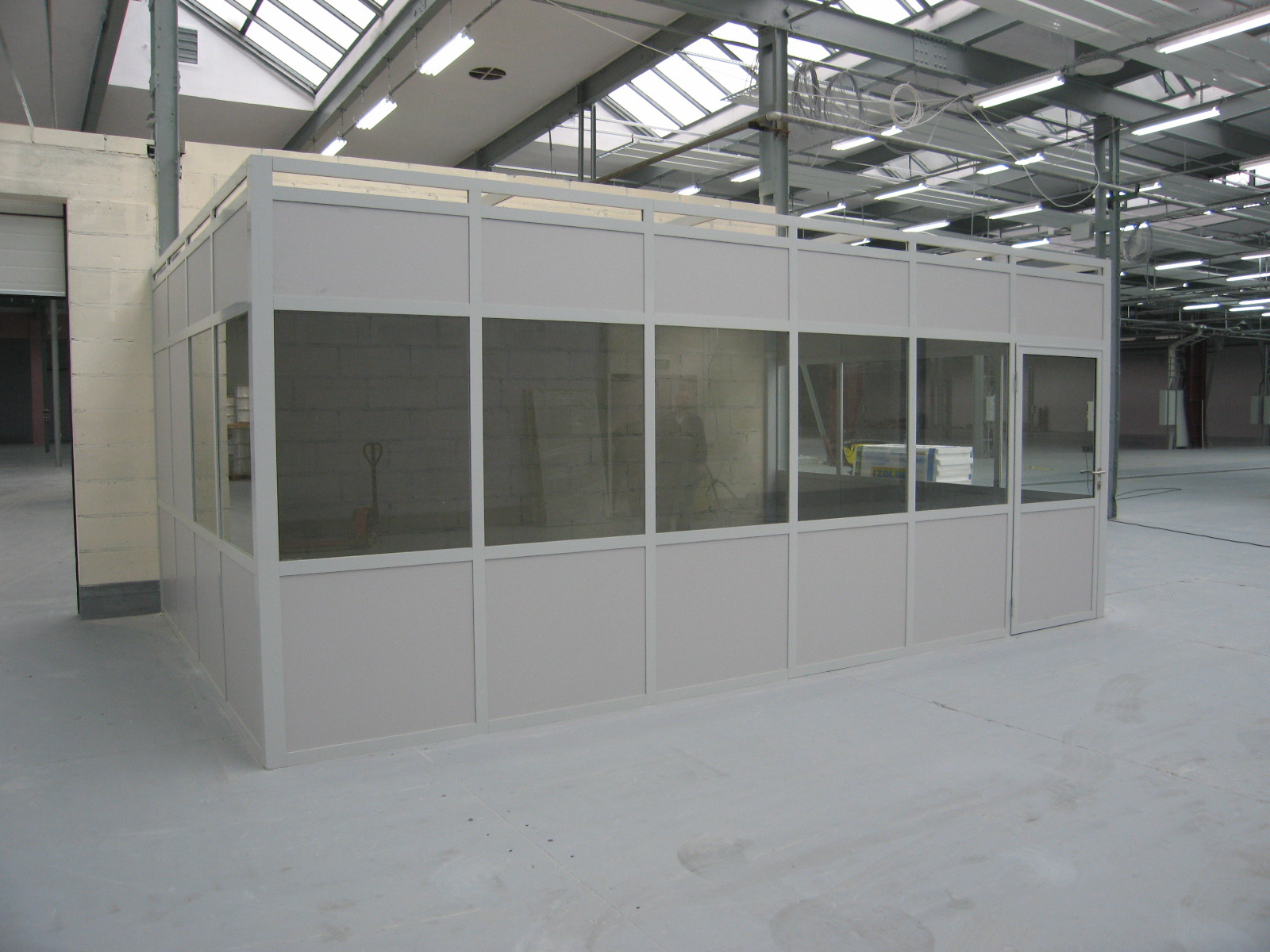 Nowoczesna, przeszklona ścianka działowa w jasnym odcieniu, oddzielająca przestrzeń w hali produkcyjnej lub magazynowej, z widocznymi elementami konstrukcyjnymi dachu i oświetleniem.
