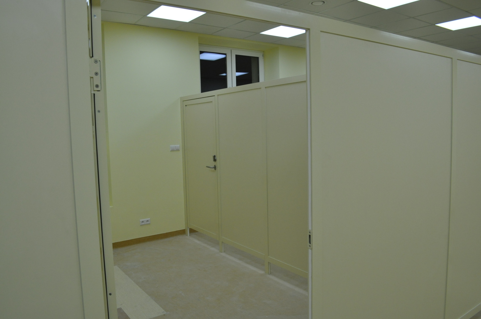 Jasne, modułowe kabiny sanitarne w biurze, widok z korytarza, ściany w odcieniach beżu, sufit z kasetonami i oświetleniem LED.