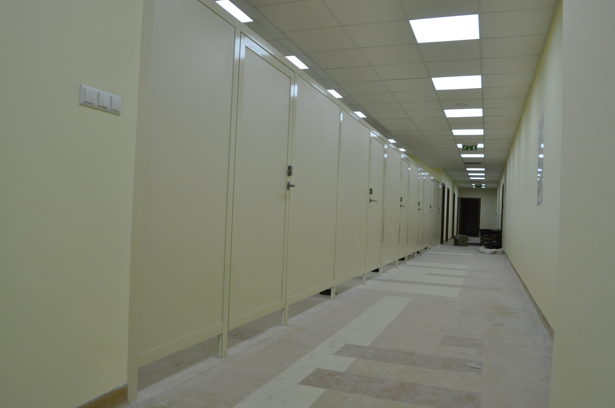 Długi korytarz z rzędem kabin sanitarnych o jasnych ścianach i podłodze w geometryczne wzory, widoczne oświetlenie sufitowe i drzwi na końcu korytarza.