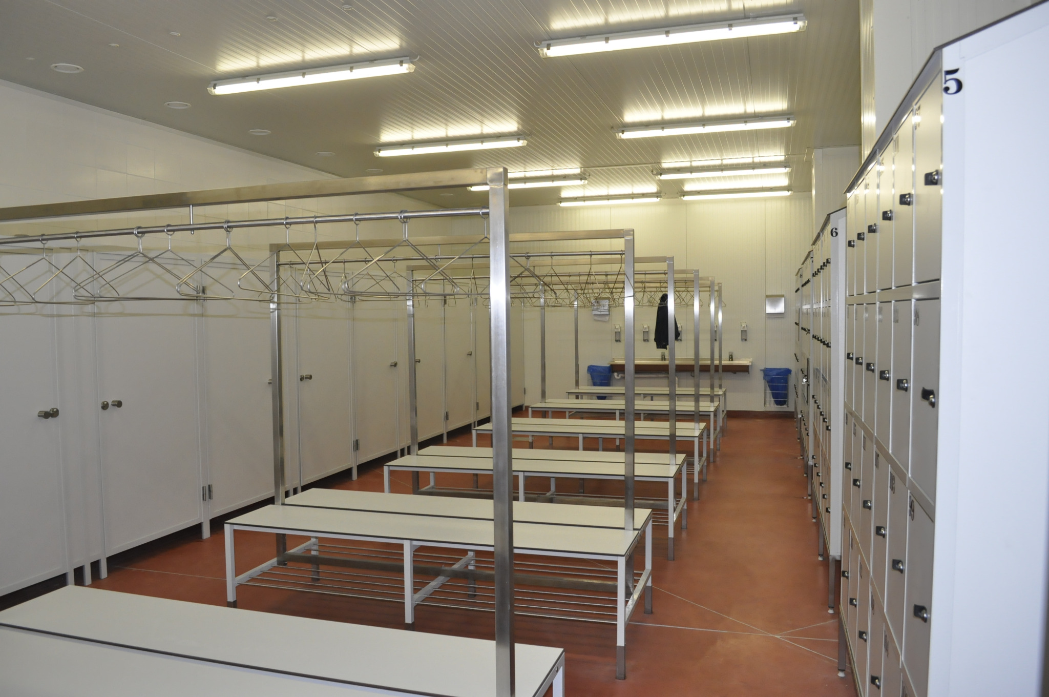 Szereg białych ławek z metalowymi stelażami i wieszakami w szatni biurowej z rzędem szafek ubraniowych z numerami, oświetlonej lampami jarzeniowymi.