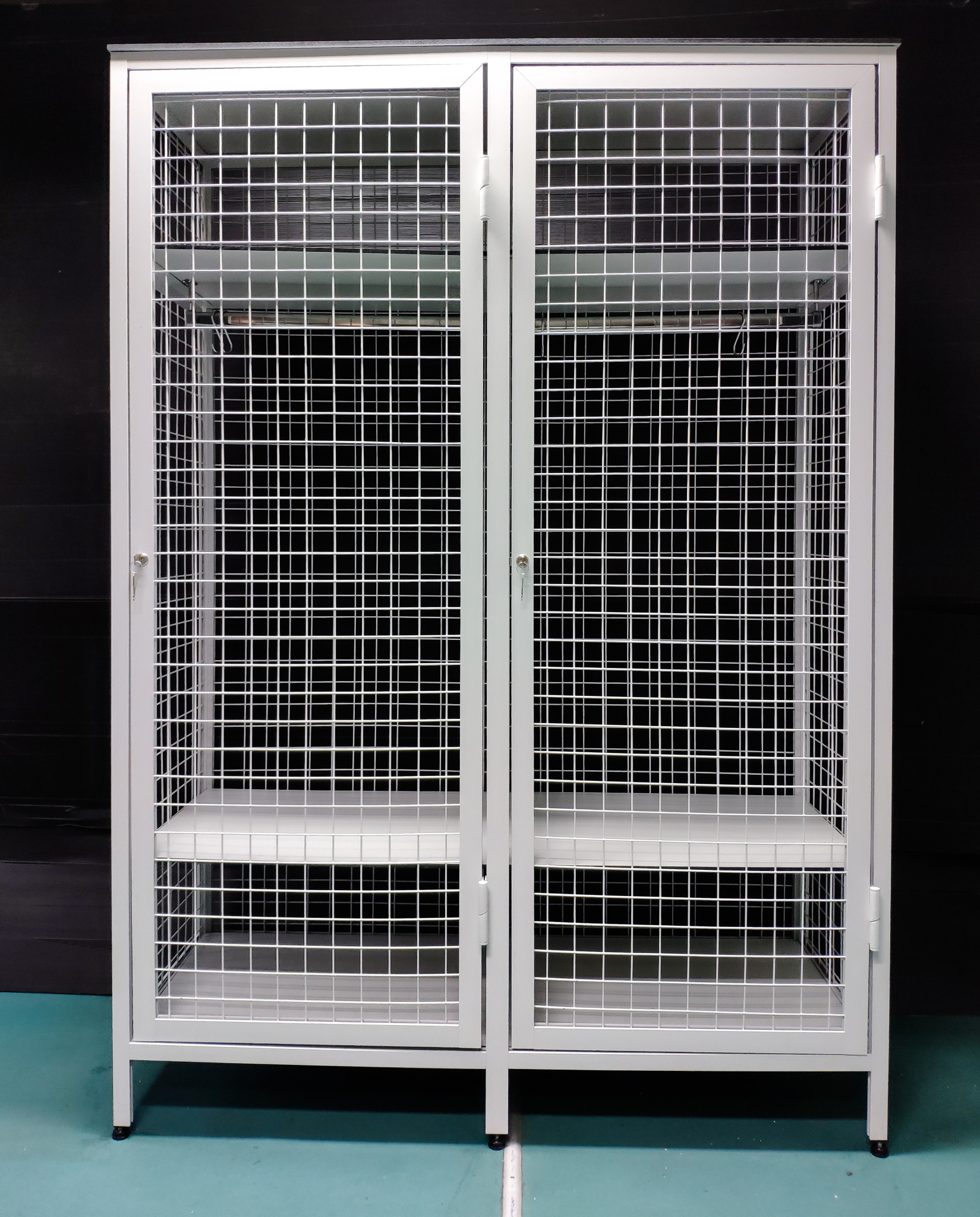 Metalowa szafa biurowa z siatki drucianej, dwudrzwiowa, zamykana na klucz, z dwoma półkami, na tle ciemnej ściany i zielonej podłogi.