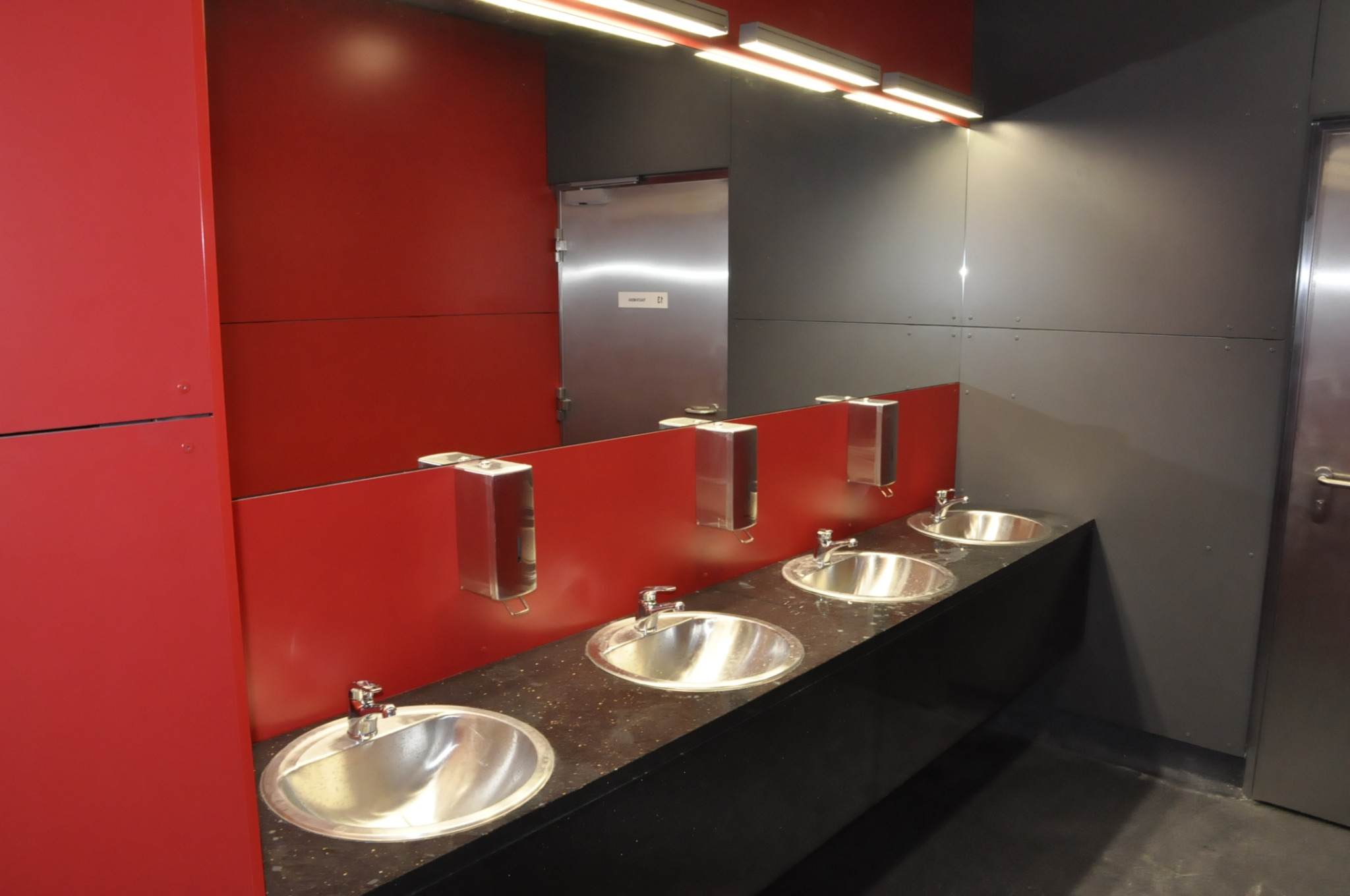 Nowoczesna łazienka biurowa z czterema umywalkami w srebrnym kolorze, czerwonymi panelami i szarymi ścianami, oświetlona listwami LED