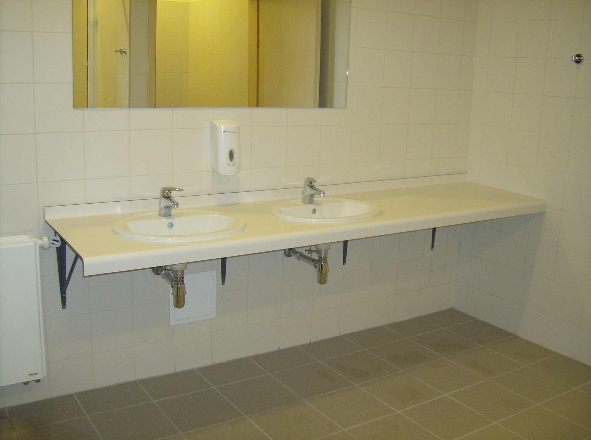 Dwie białe umywalki zamontowane na wspólnym blacie w toalecie biurowej, z widocznymi chromowanymi kranami, dozownikiem mydła na ścianie i szarą podłogą z płytek.