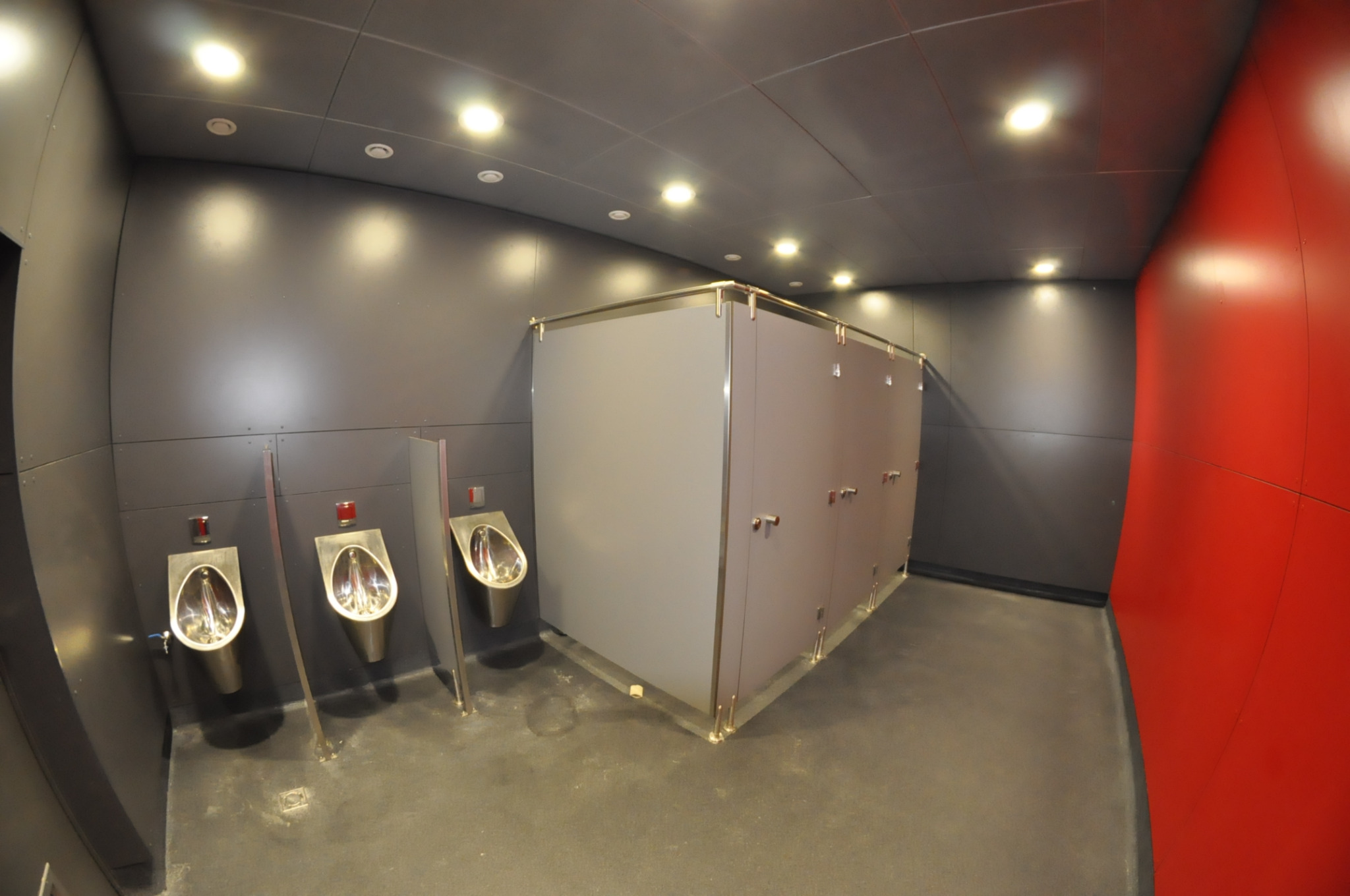 Nowoczesna toaleta publiczna w biurze: szare ściany, rzędy metalowych pisuarów z czerwonymi detalami i kabiny WC z metalowymi obramowaniami, oświetlenie sufitowe