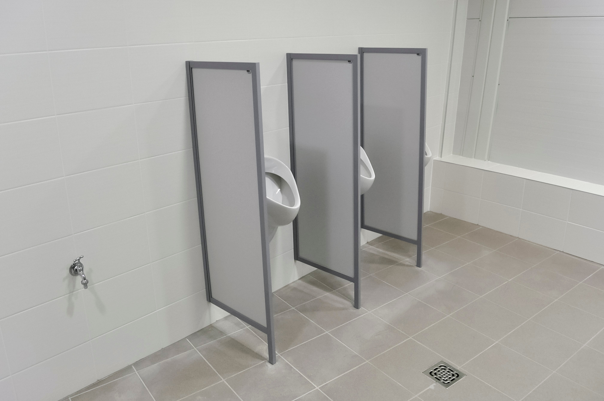 Nowoczesna toaleta biurowa w Piastowie z trzema pisuarami oddzielonymi szarymi ściankami działowymi, wykończona białymi płytkami i szarą podłogą z kratką ściekową.