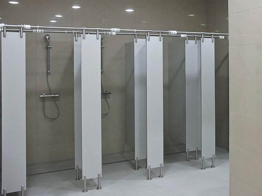 Rząd kabin prysznicowych z szarymi ściankami i stalowymi nogami w jasnej łazience z szarymi płytkami ściennymi i podłogowymi, widoczne baterie prysznicowe.