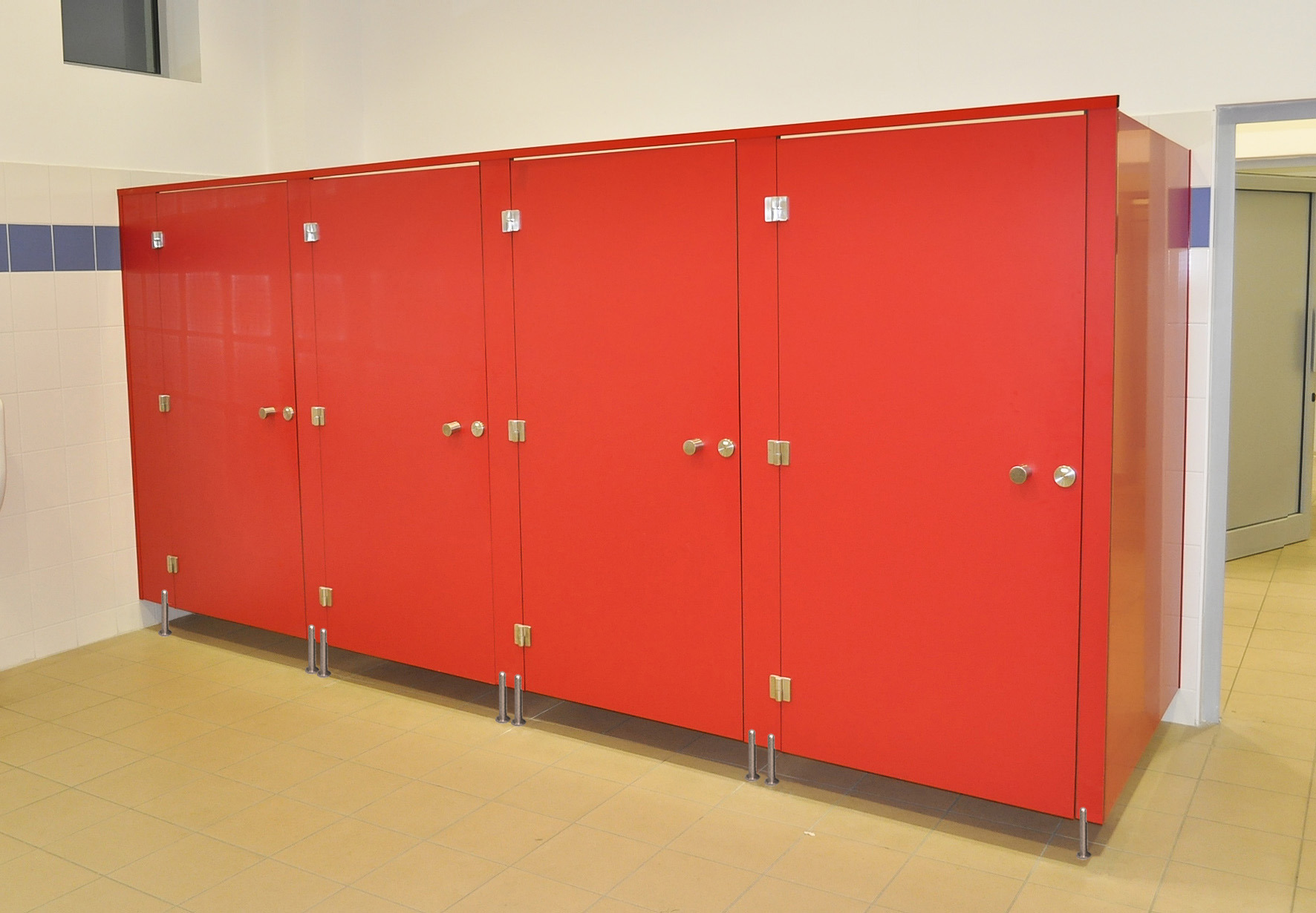 Czerwone kabiny toaletowe z metalowymi okuciami w biurowej łazience, widoczne płytki na ścianach i podłodze.
