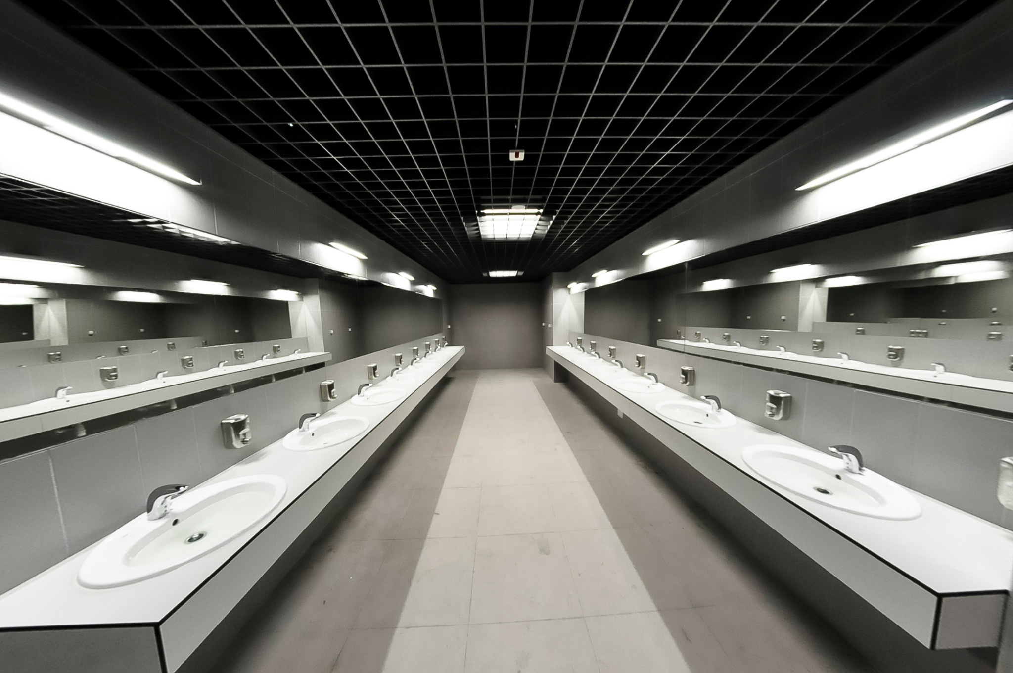 Długi rząd umywalek w biurowej toalecie z lustrami, czarną kratownicą sufitową i jasnym oświetleniem.