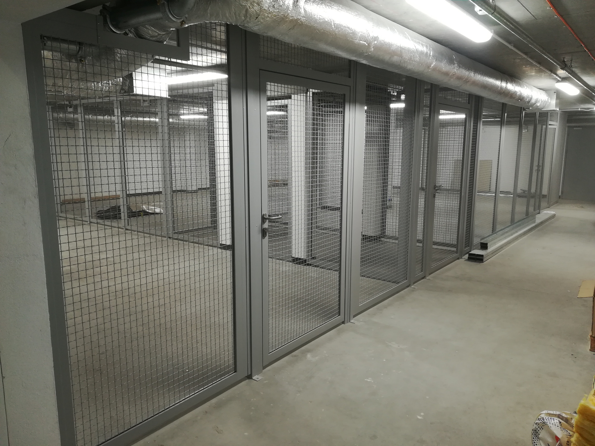 Klatki magazynowe z siatki w szarych ramach, rzędem wzdłuż korytarza biurowego, widoczne rury wentylacyjne pod sufitem.