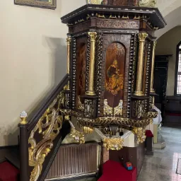 Bogato zdobiona ambona w kościele, z drewnianymi panelami z malowidłami i złoceniami, widoczne schody z rzeźbioną balustradą i obraz Matki Boskiej w tle.