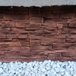 Dekoracyjna ściana z paneli imitujących czerwoną cegłę, wykończona białymi otoczakami u podstawy i szarym tynkiem na górze.