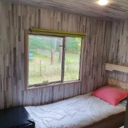 Pokój w domku letniskowym z łóżkiem jednoosobowym, oknem z widokiem na zieleń i ścianami wyłożonymi panelami imitującymi drewno.