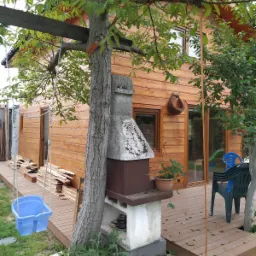 Drewniany dom z tarasem, na którym stoi murowany grill z kominem obrośniętym mchem, obok huśtawka dla dziecka zawieszona na drzewie, a w tle ułożone deski.