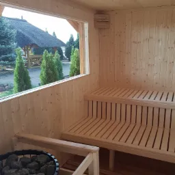 Wnętrze sauny z drewnianymi ławkami i piecem na kamienie, widok na zewnątrz przez okno na ogród z altaną krytą strzechą.