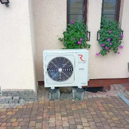 Biała pompa ciepła Rotenso Aquami zamontowana na betonowych bloczkach przed budynkiem z dwoma oknami ozdobionymi skrzynkami z fioletowymi kwiatami, obok stos kostki brukowej.