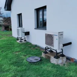 Dwa zewnętrzne moduły klimatyzacji Panasonic zamontowane na wspornikach przy ścianie domu z szarą stolarką okienną, widoczny trawnik i studzienka kanalizacyjna.