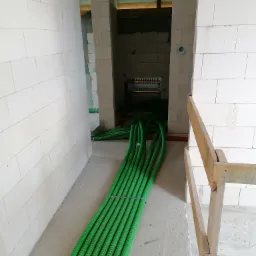 Wiązka zielonych rur karbowanych ułożona na surowej posadzce w trakcie montażu instalacji w budynku z białego bloczka, widoczny rozdzielacz w tle.
