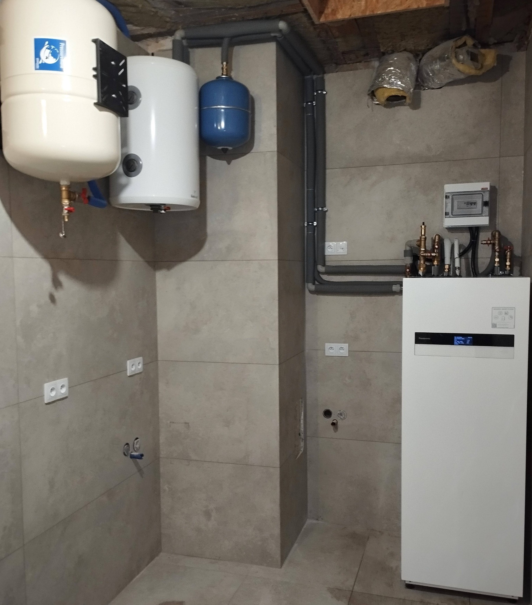 Instalacja z białym zbiornikiem ciśnieniowym z logo, białym bojlerem i niebieskim naczyniem przeponowym, szare rury rozprowadzone po ścianie z gniazdkami elektrycznymi oraz biała jednostka pompy...