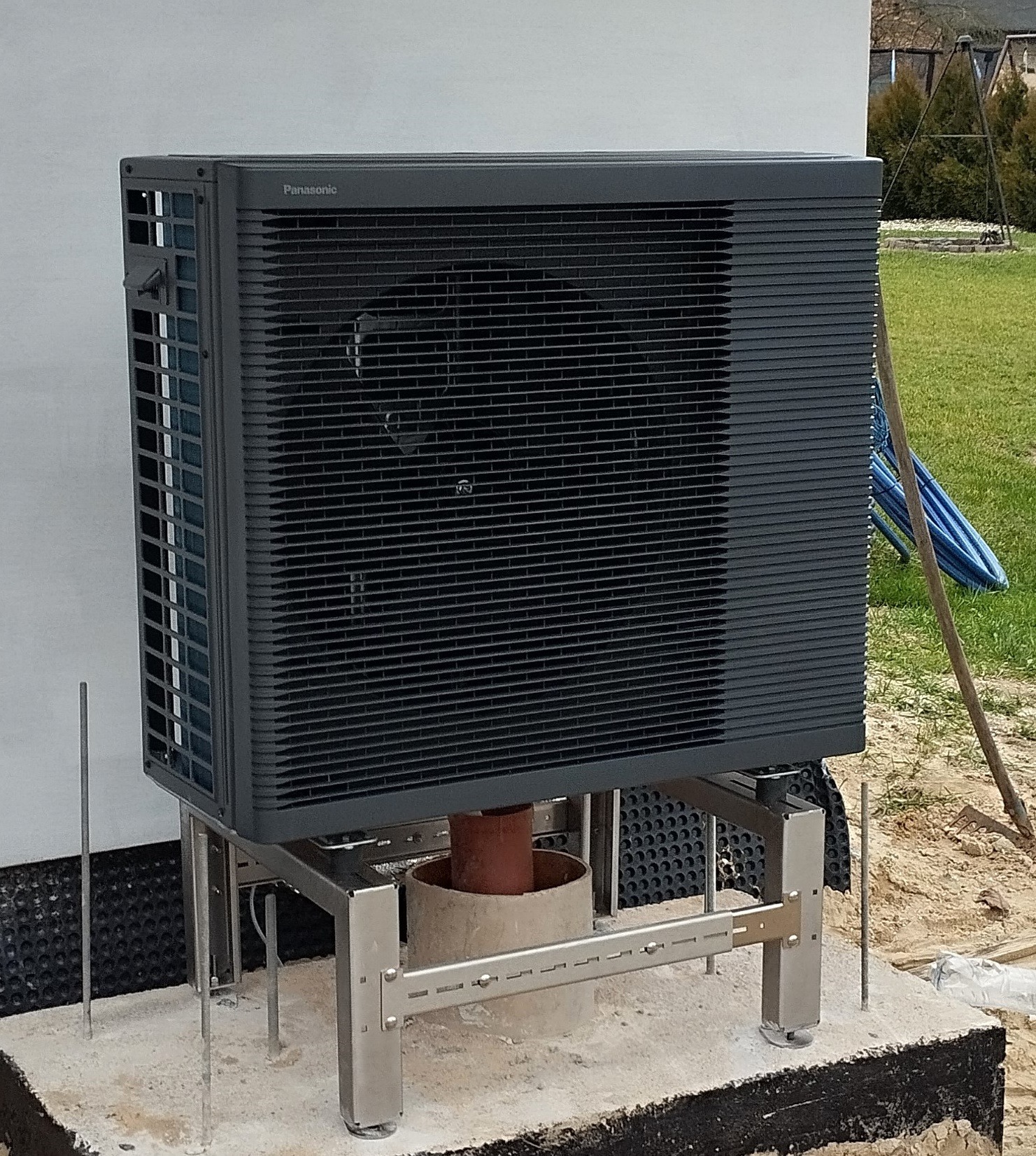Zewnętrzna jednostka pompy ciepła Panasonic zamontowana na metalowej konstrukcji na betonowym fundamencie, widoczne elementy instalacji i otoczenie budynku.