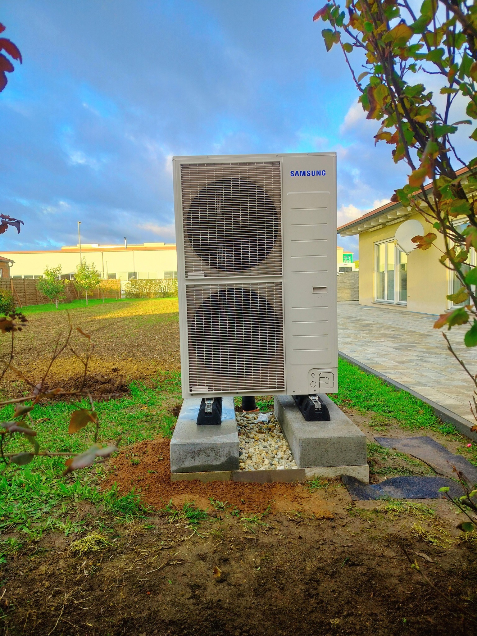 Biała, dwusekcyjna pompa ciepła marki Samsung, zamontowana na betonowej podstawie z drobnymi kamieniami, na tle trawnika i częściowo zarośniętego ogrodu w pochmurny dzień.