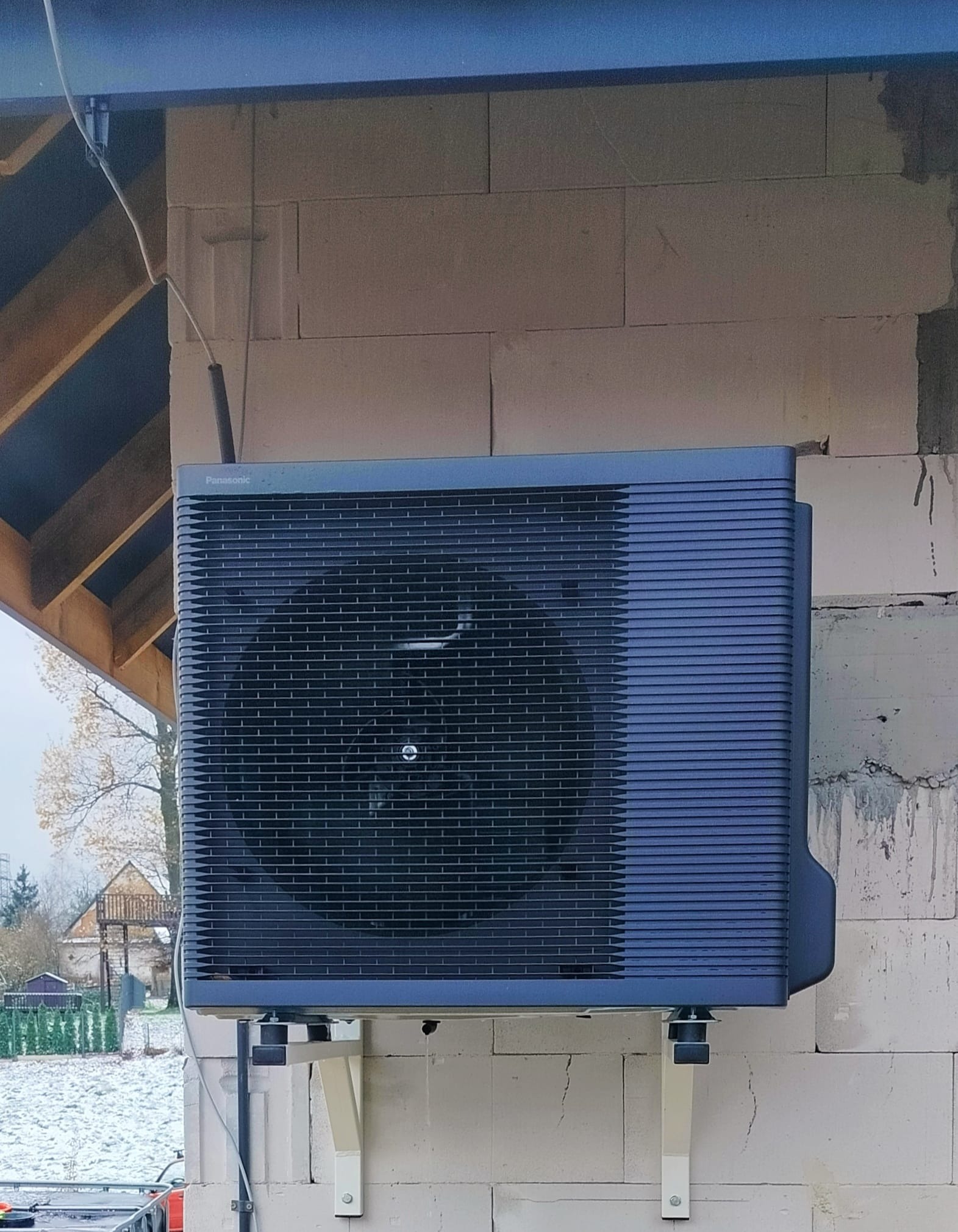 Zewnętrzna jednostka pompy ciepła Panasonic zamontowana na wspornikach przy ścianie budynku z bloczków, widoczny fragment konstrukcji dachu i zaśnieżony krajobraz w tle.