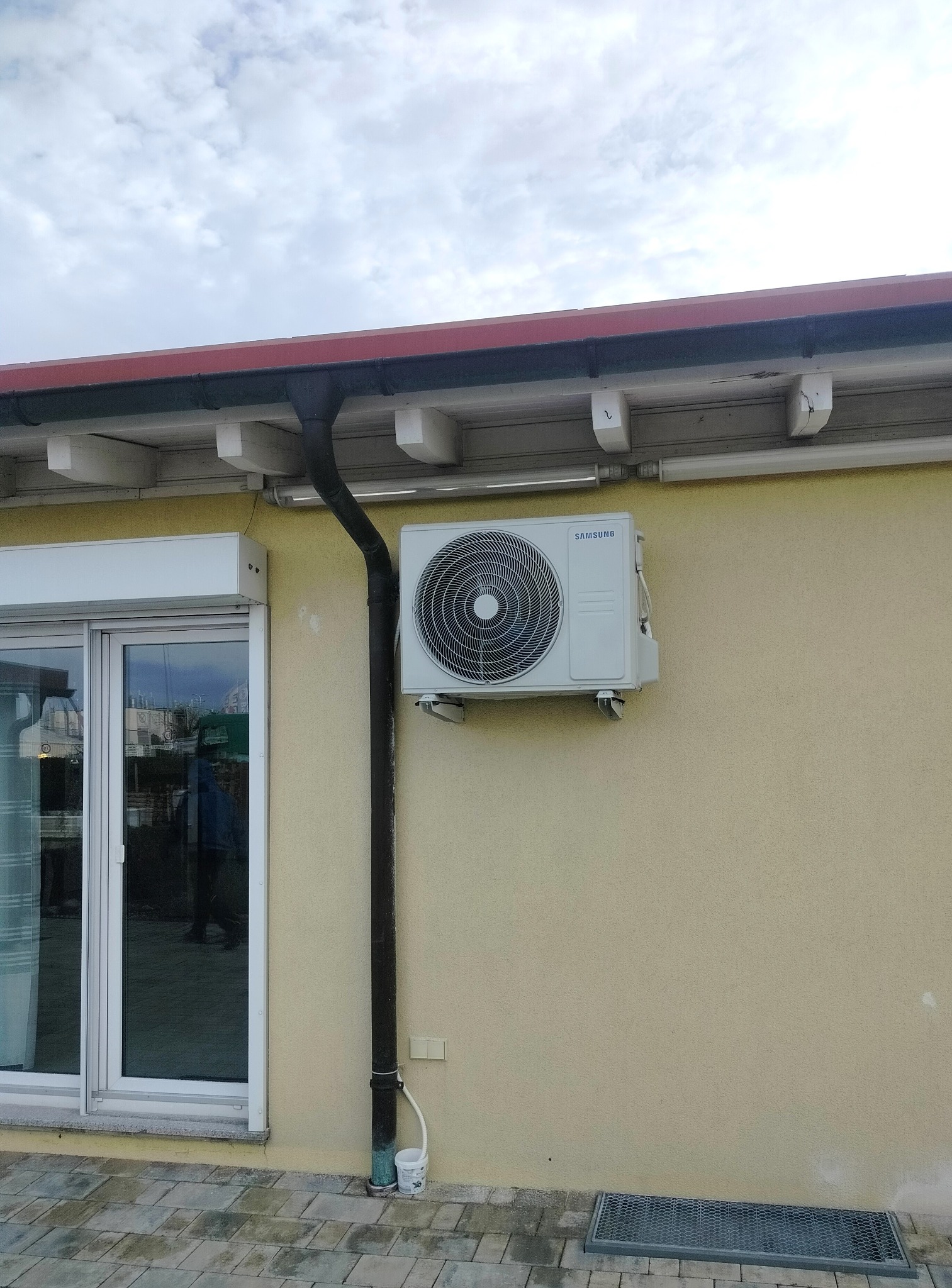 Zewnętrzna jednostka klimatyzacji Samsung zamontowana na beżowej ścianie budynku, obok rynny spustowej i okna z roletą.