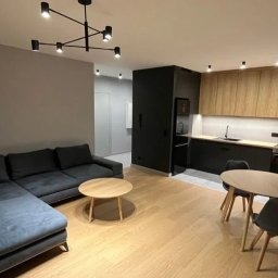 Nowoczesne wnętrze mieszkania z szarą sofą narożną, okrągłym drewnianym stolikiem kawowym, drewnianą podłogą, czarną zabudową kuchni z drewnianymi szafkami i okrągłym stołem jadalnym z szarymi...