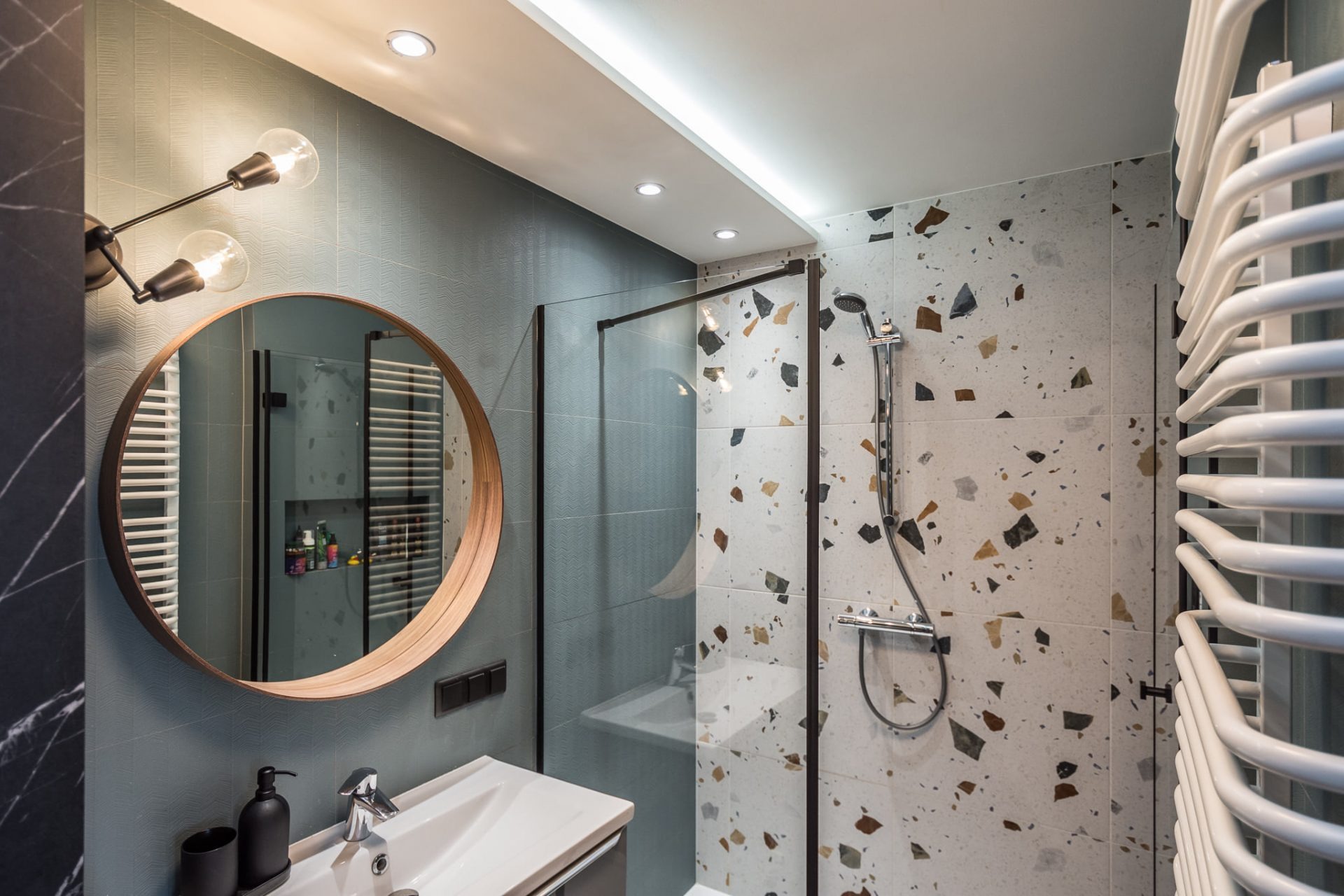 Nowoczesna łazienka z okrągłym lustrem z drewnianą ramą, kabiną prysznicową ze szklaną ścianką i mozaikową ścianą, oświetlona lampą z żarówkami Edisona i ledowym paskiem na suficie.