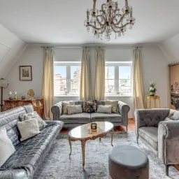 Salon w mansardzie z szarymi sofami, marmurowym stolikiem kawowym na złotych nogach, kryształowym żyrandolem i widokiem na starówkę przez okna.