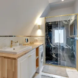 Jasna łazienka na poddaszu z marmurowymi ścianami, drewnianym blatem pod umywalką i kabiną prysznicową z czarnym marmurem oraz złotymi akcentami.