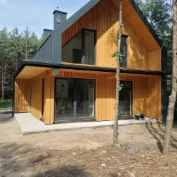 Nowoczesny dom jednorodzinny z drewnianą elewacją i ciemnoszarym dachem, widok z zewnątrz w słoneczny dzień, na pierwszym planie młode drzewko.