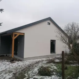 Nowoczesny dom jednorodzinny z szarym dachem i drewnianą wiatą przy wejściu, częściowo ośnieżony teren.