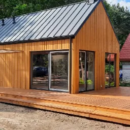 Nowoczesny, parterowy dom z drewnianą elewacją, antracytowym dachem z blachy na rąbek stojący i przestronnym tarasem z drewna, otoczony zielenią.