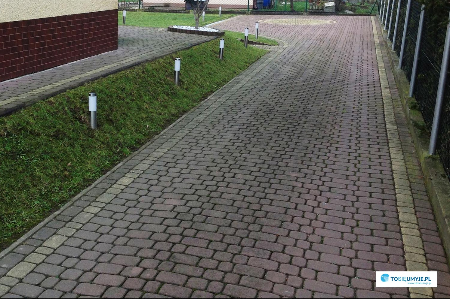 Ciemnoczerwona kostka brukowa ułożona na podjeździe z oświetleniem w postaci niskich lamp po obu stronach, obok trawnika i ogrodzenia.