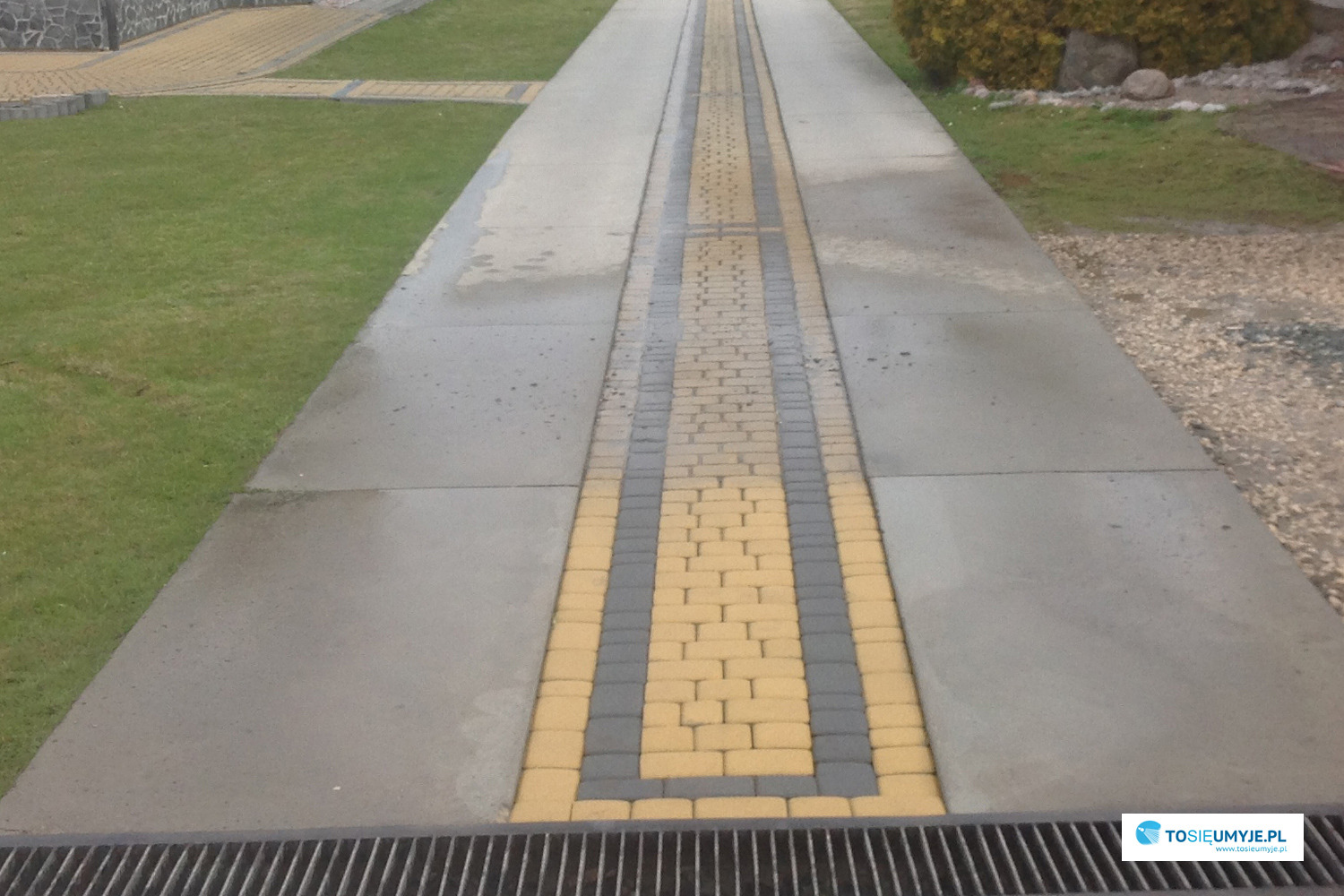 Długa, prosta ścieżka z kostki brukowej w odcieniach szarości i żółci, otoczona trawnikiem, widziana z perspektywy lekko z góry, zakończona metalową kratką ściekową.