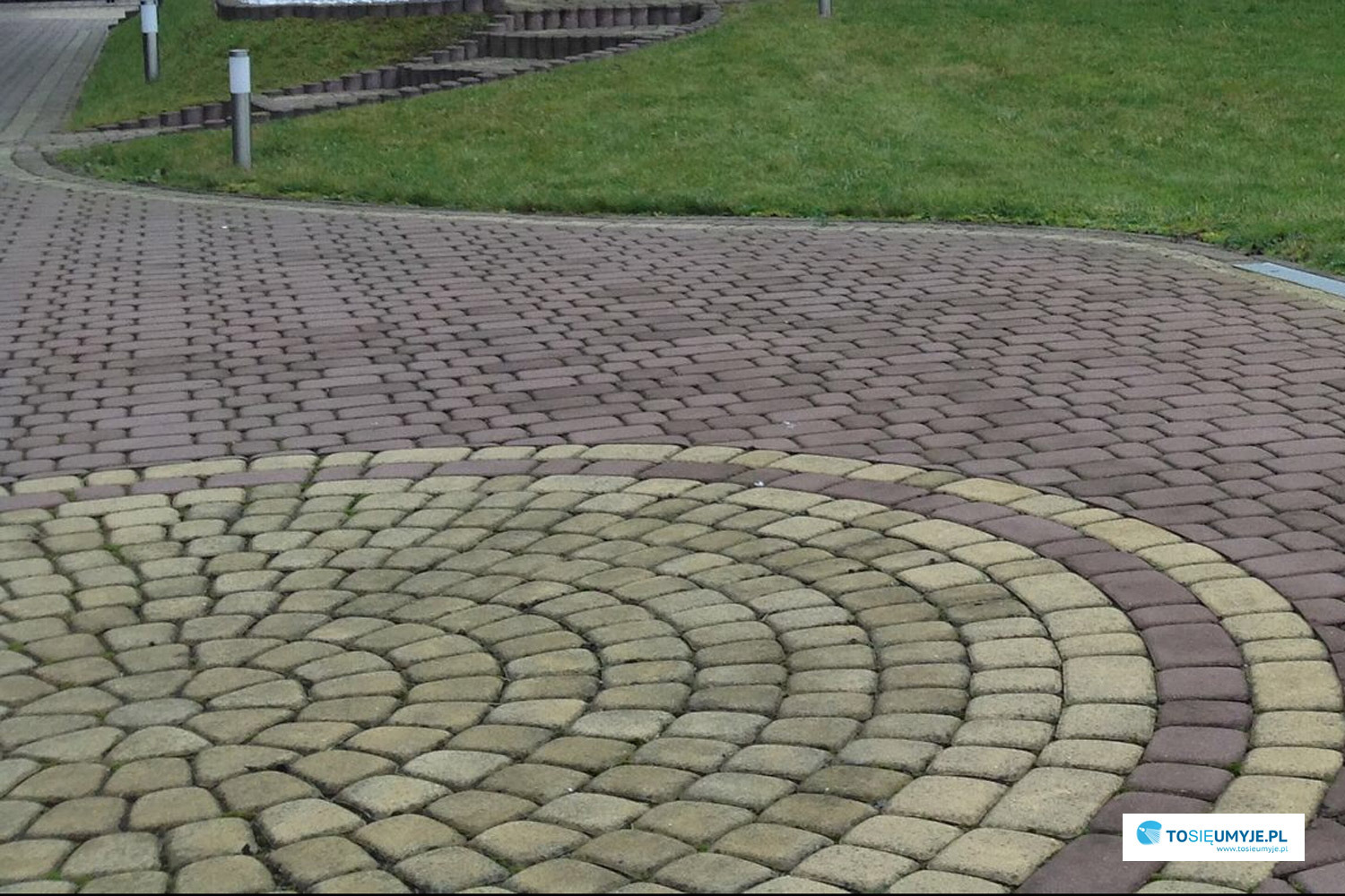 Ułożona kostka brukowa w dwóch kolorach: brązowym i beżowym, tworząca wzór koła na podjeździe, z trawnikiem i schodami w tle.