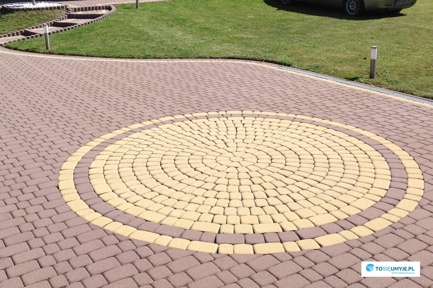 Ułożona kostka brukowa w odcieniach brązu i beżu tworząca okrągły wzór na podjeździe, widoczne schody i trawnik w tle.
