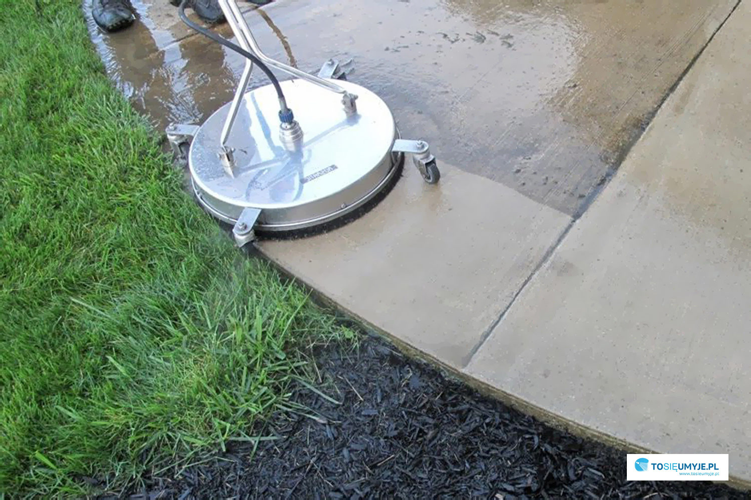 Czyszczenie kostki brukowej urządzeniem rotacyjnym z dyszami, widoczna różnica w czystości między umytą a brudną powierzchnią, trawa i czarne kamyki w tle.