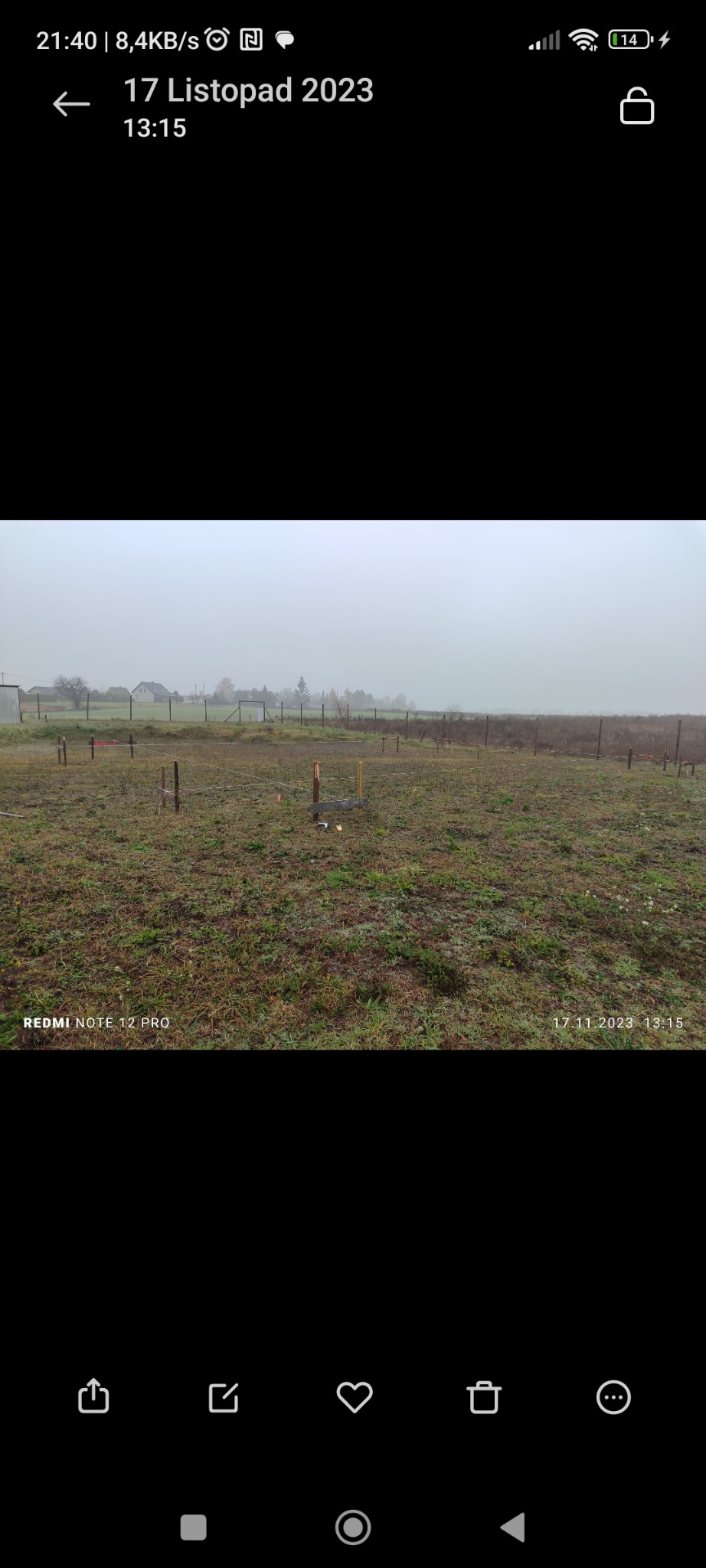 Wytyczanie działki w terenie: drewniane paliki wbite w ziemię, wyznaczające granice. Mglista pogoda, widoczne domy w oddali. Prace pomiarowe w toku.