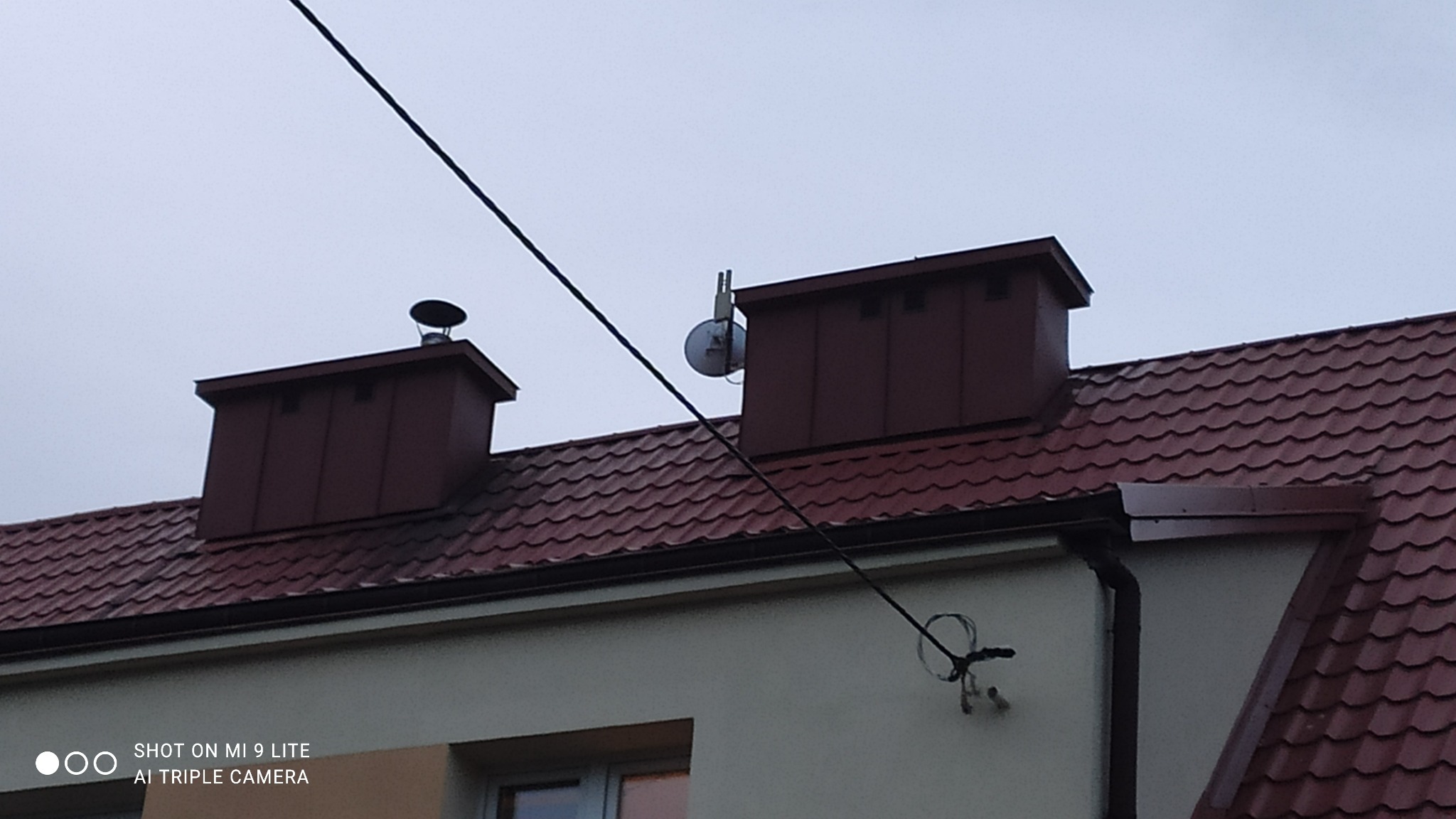 Czerwony dach z dachówki ceramicznej z dwoma kominami i anteną satelitarną, widoczne przewody elektryczne na tle pochmurnego nieba.