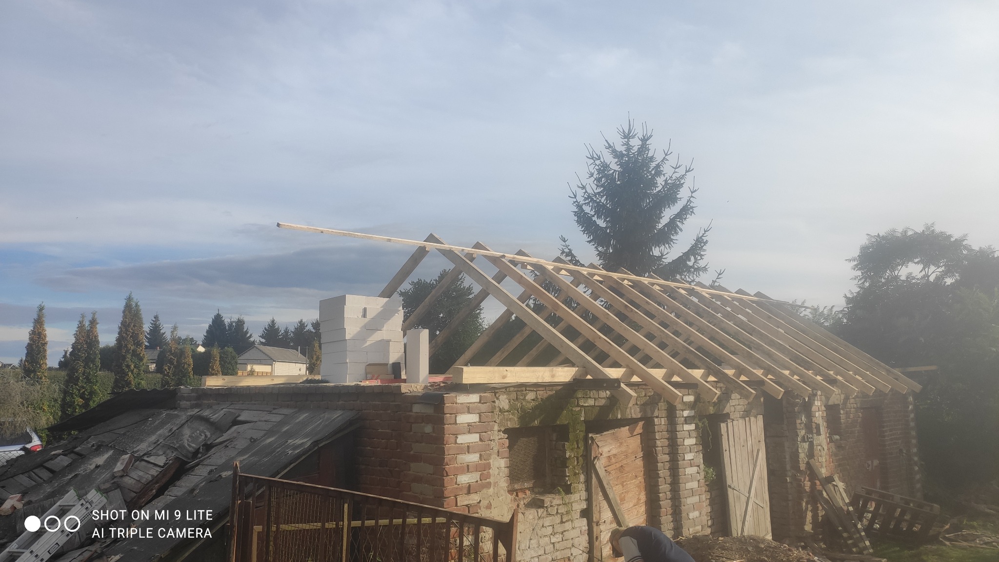 Konstrukcja drewniana więźby dachowej na budynku z cegły, widoczne bloczki betonowe i stara papa na dachu, drzewa w tle, pochmurne niebo.