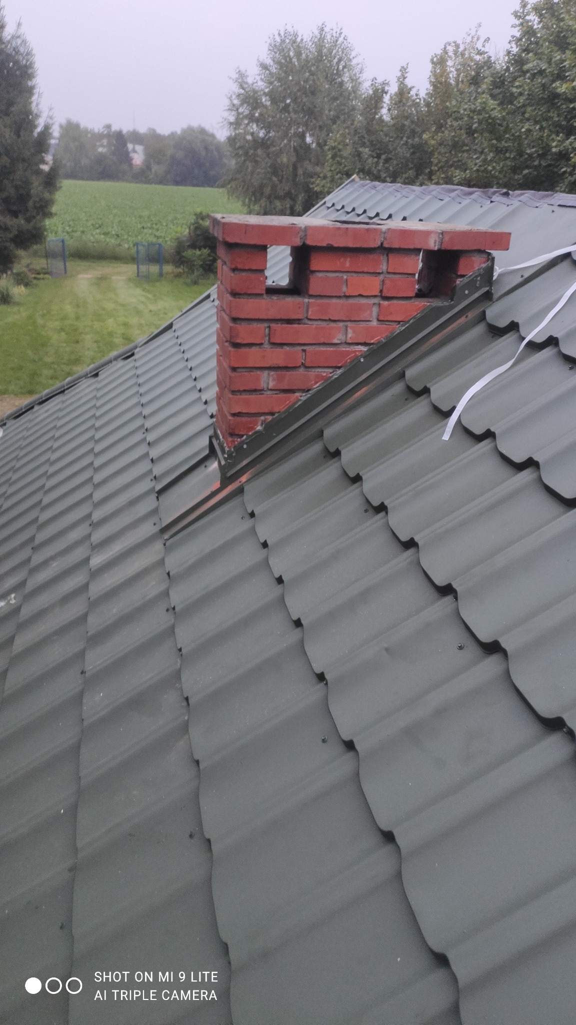 Nowy, zielony dach z blachy trapezowej z czerwonym kominem z cegły, widok z góry na tle zielonego krajobrazu.