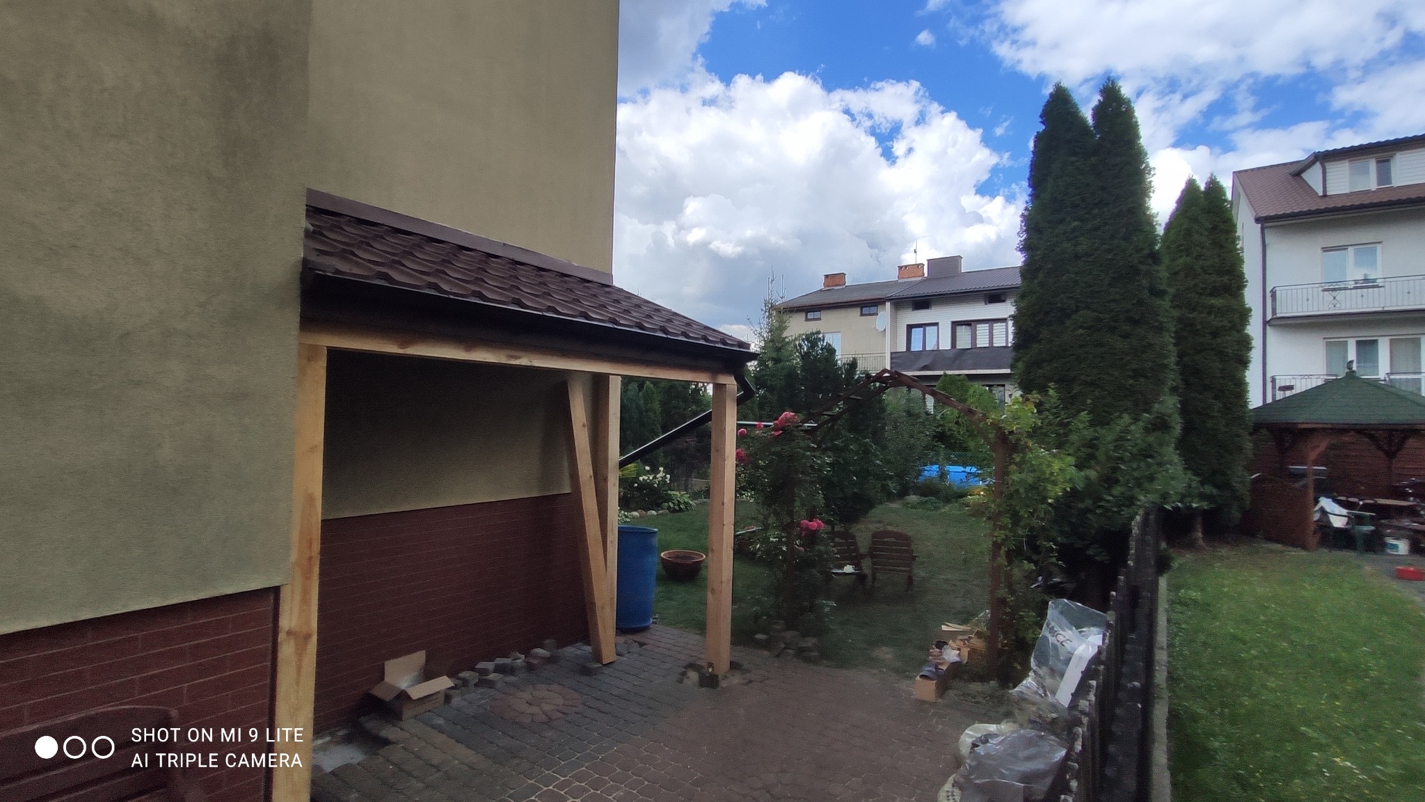 Zadaszenie tarasu w trakcie budowy, widoczne drewniane słupy nośne i konstrukcja dachu pokryta brązową blachodachówką, w tle ogród z zielenią i zabudowaniami.