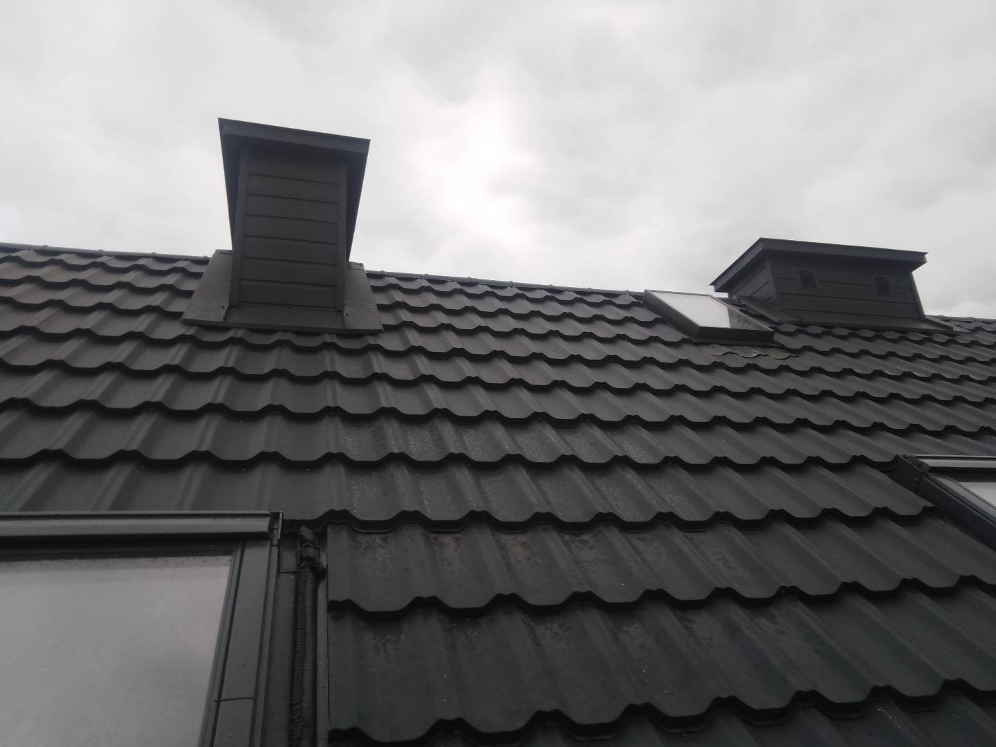 Czarny dach z blachodachówki z dwoma kominami wentylacyjnymi i oknem dachowym, widok pod kątem z dołu, pochmurne niebo w tle.