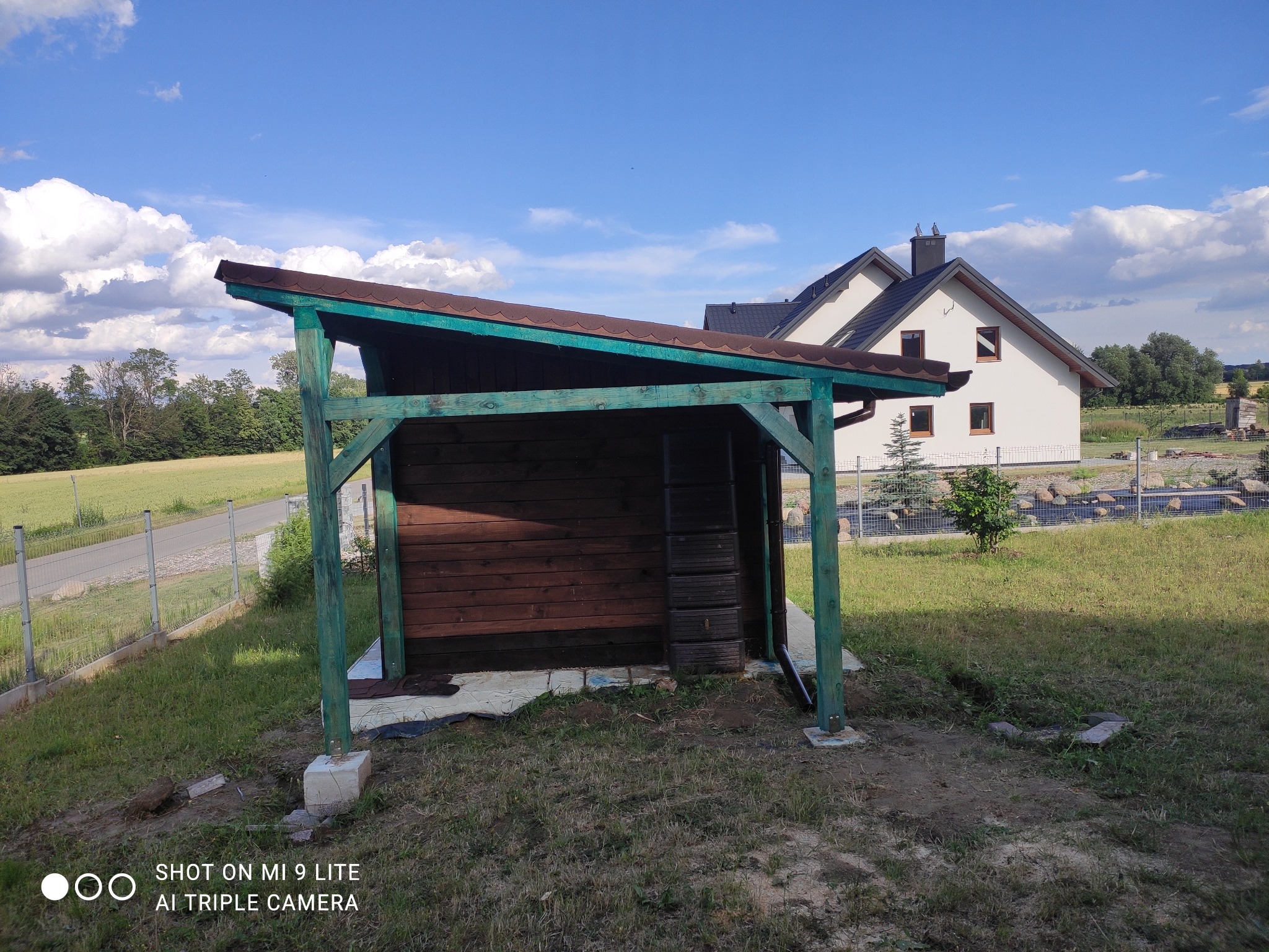 Zadaszona drewniana wiata z brązowym dachem i zielonymi słupami, na trawiastym terenie, z domem w tle.