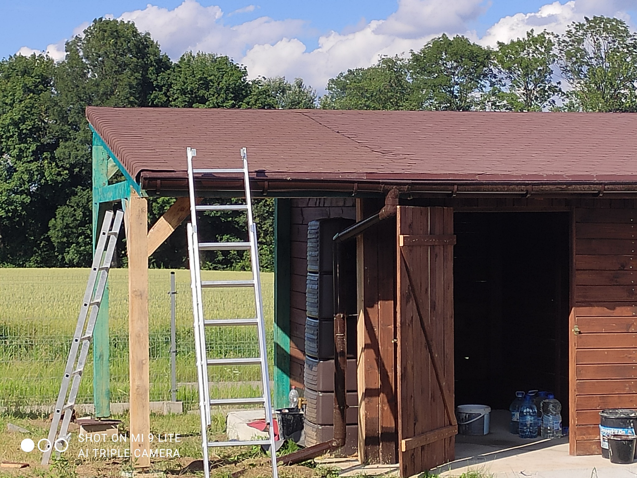 Brązowy dach z gontów bitumicznych na drewnianej konstrukcji, widoczna drabina oparta o budynek gospodarczy z otwartymi drzwiami, w tle pole i drzewa.