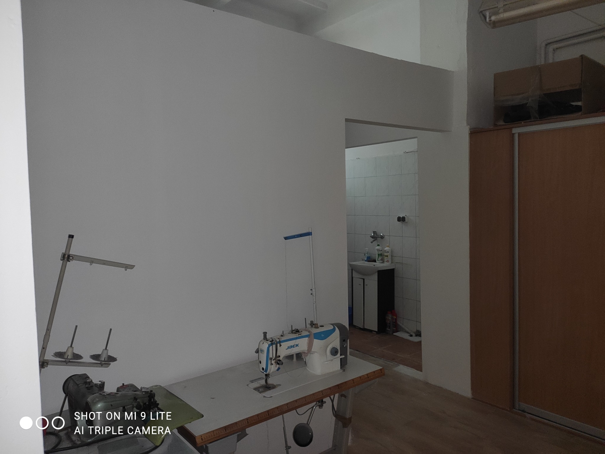 Wnętrze pomieszczenia z dwiema maszynami do szycia, jedna starsza, druga nowsza, na tle białych ścian i widocznej łazienki w głębi.