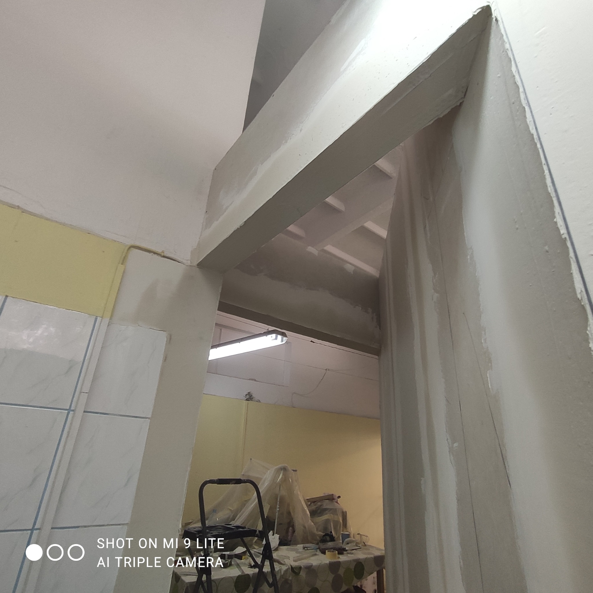 Przygotowanie do malowania futryny drzwiowej, widoczna drabina i stół z narzędziami malarskimi, ślady szpachlowania na ścianach i suficie.