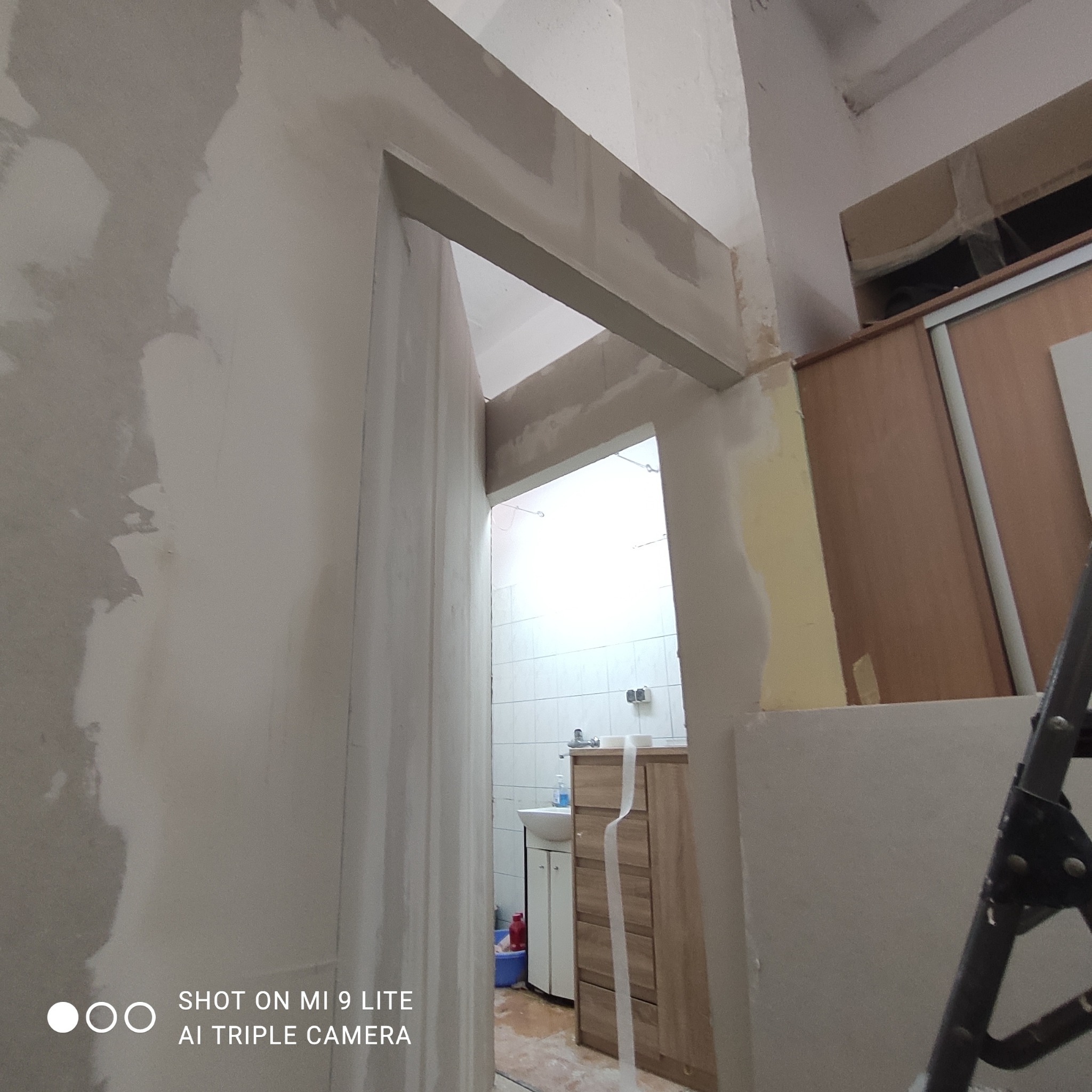 Przygotowanie do malowania: ściana z widocznymi śladami szpachlowania, surowy otwór drzwiowy, fragment łazienki w tle z widocznym umywalką i szafką, drabina oparta o ścianę po prawej stronie kadru.